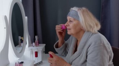 Yaşlı büyük anne cilt bakımı yapıyor, kırışıklık önleyici temeli süngerle uyguluyor.