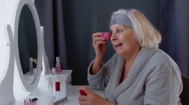 Yaşlı büyük anne makyaj yapıyor, kırışıklık önleyici temeli süngerle uyguluyor.