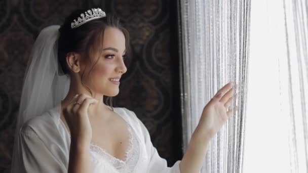 Novia en vestido de tocador cerca de la ventana. Preparativos matutinos. Mujer con vestido de noche y velo — Vídeos de Stock