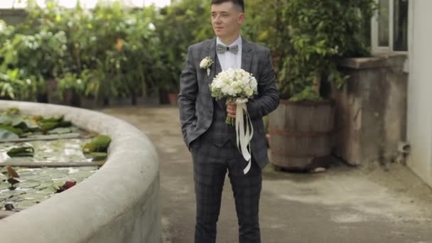 Brudgummen går ner i gränden mellan buskar med en bröllopsbukett till sin älskade brud — Stockvideo
