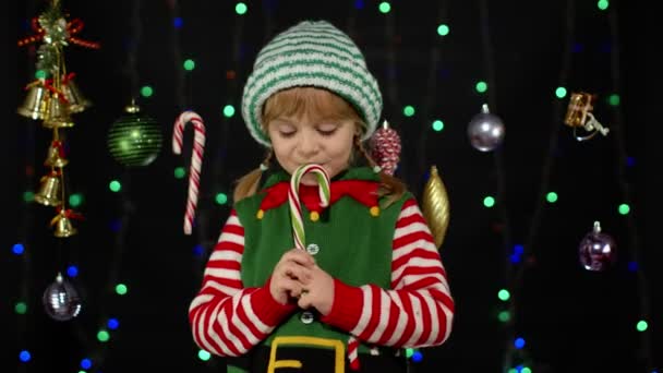 Niña en Navidad elfo Santa Claus ayudante traje lamiendo caramelo bastón piruleta caramelo dulces — Vídeos de Stock