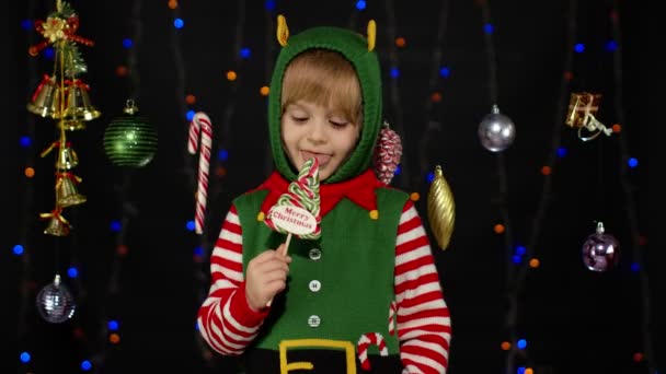 Kid girl in Christmas elf Santa Claus helper costume licking candy lollipop caramel sweets — Stock Video