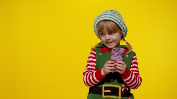 Niña con teléfono móvil en Navidad elfo Santa ayudante traje señala los dedos a un lado en la pared en blanco — Vídeo de stock