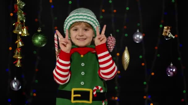 Blondýny dítě dívka v vánoční skřítek Santa Claus helper kostým dělat mír gesto, ukazuje v znamení — Stock video