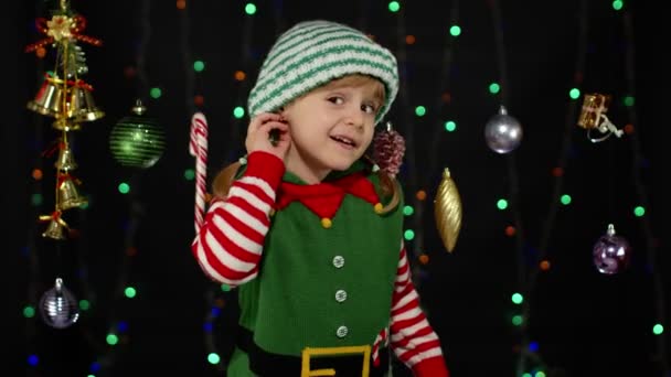 Chica en traje de Navidad mantiene la mano cerca de la oreja trata de escuchar lo que los padres actuales van a dar — Vídeo de stock