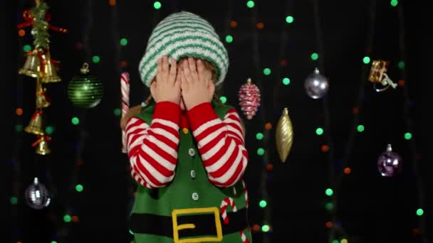 Kid girl in Christmas elf Santa Claus helper costume cover face with hands and plays hide and seek — Stock Video
