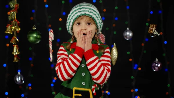 Menina criança no elfo de Natal Papai Noel ajudante traje olha para a câmera faz grandes olhos cobrindo boca — Fotografia de Stock