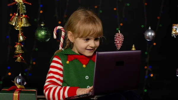 Kid girl in Christmas elf costume doing shopping online using laptop, browsing on social media
