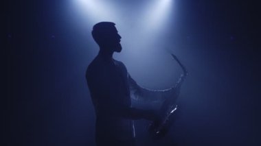 Karanlık gece kulübü stüdyosunda saksafoncu bir adamın arka ışıklandırılmış silueti ve saksafon çalmaya başla.