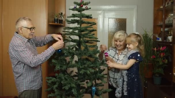 Children girl with elderly couple grandparents decorating artificial Christmas pine tree at home — Stock Video