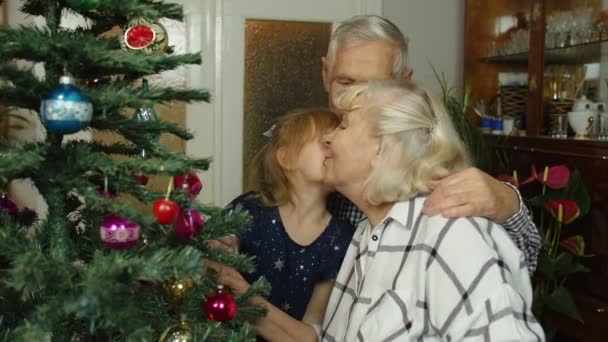 Маленькая милая девочка со старшими бабушкой и дедушкой украшают искусственную елку дома — стоковое видео