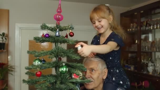 Niña con abuelo mayor decorando árbol de Navidad artificial con adornos y juguetes en casa — Vídeos de Stock