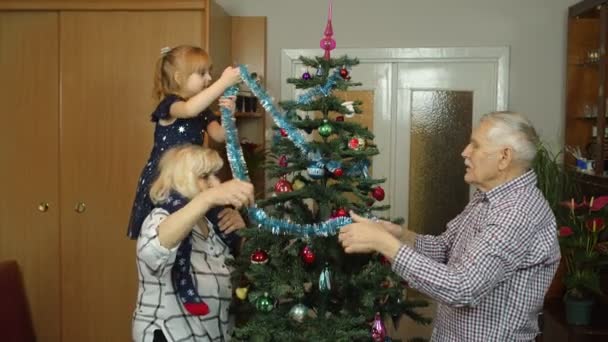 Kindermädchen mit betagten Großeltern schmücken künstlichen Tannenbaum zu Hause — Stockvideo