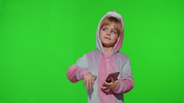 Κορίτσι παιδί σε μονόκερο πιτζάμα χρησιμοποιώντας, κρατώντας smartphone, δείχνοντας κάτι με τα χέρια, αντίγραφο χώρο — Αρχείο Βίντεο