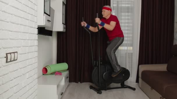 Hombre mayor en ropa deportiva usando orbitrek en la sala de estar haciendo ejercicios de entrenamiento deportivo de cardio en casa — Vídeos de Stock