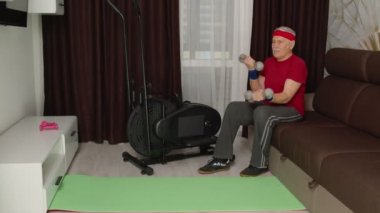 Spor giyim bölümünde yaşlı bir adam ağırlık kaldırıyor. Halter kaldırıyor. Evde egzersiz yapıyor.