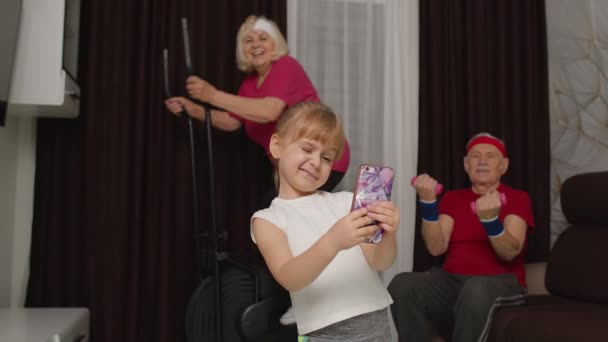 Casal sênior usando orbitrek fazendo exercícios de halteres, criança menina fazendo selfie no telefone móvel — Vídeo de Stock