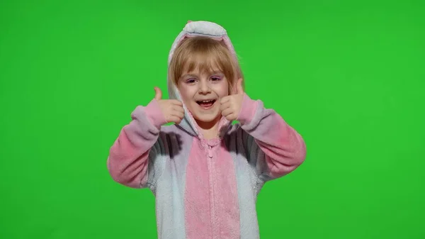 Mała dziewczynka uśmiecha się, pokazując kciuki w górę gest, zgadzają się znak w piżamie jednorożca na klawiszu chroma — Zdjęcie stockowe