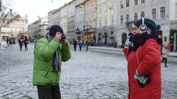 Idősebb feleség pózol a kamera előtt nevetve, miközben a férj képeket készít a Winter City utcában. — Stock Fotó