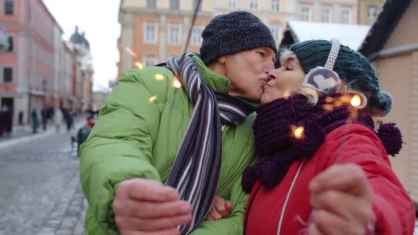 Starší pár drží jiskřičky bengálské světla těší Štědrý večer, dělat polibek v zimním městě — Stock video