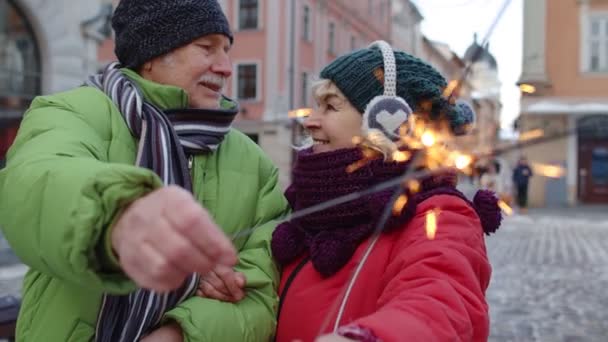 Ηλικιωμένο ζευγάρι κρατώντας sparklers φώτα bengal απολαμβάνοντας την παραμονή των Χριστουγέννων, κάνοντας φιλί στη χειμερινή πόλη — Αρχείο Βίντεο