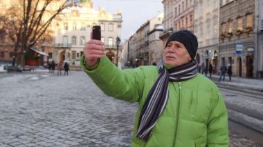 Kış şehir merkezinde kıdemli turist selfie çekiyor, akıllı telefonuyla online video görüşmesi yapıyor.