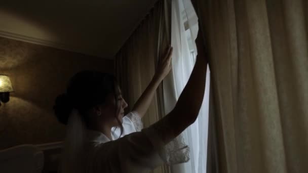 Noiva em vestido boudoir branco ficar perto da janela e cortinas de abertura, mulher de vestido de noite e véu — Vídeo de Stock