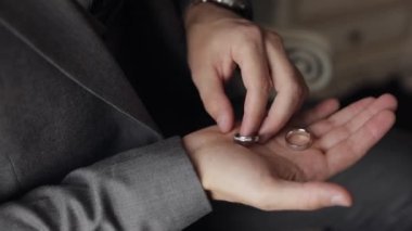 Gri ceketli damat avucunun içinde alyansları tutuyor, adam yüzüklere dokunuyor, ağır çekimde.