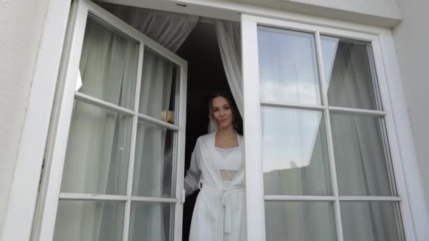 Mariée en robe de boudoir blanc va au balcon et ouvre les rideaux et la porte dans l'appartement de la chambre d'hôtel — Video