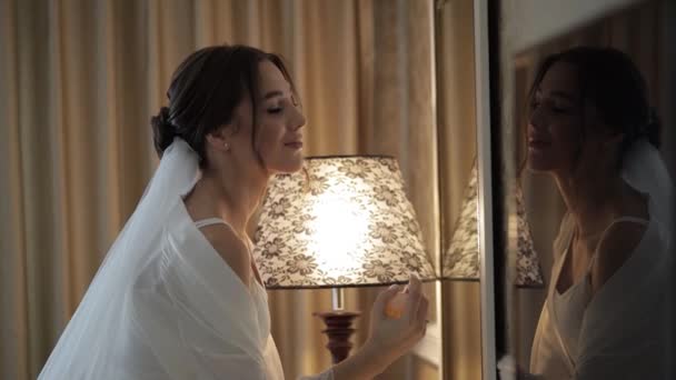 Noiva em vestido boudoir branco ficar perto de espelho olhando para reflexão, usando perfume ou desodorizante — Vídeo de Stock