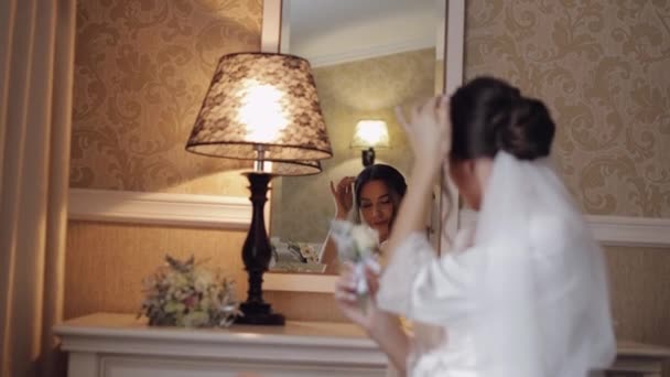 Novia en vestido de tocador blanco sentado cerca del espejo mirando a la reflexión, los preparativos de la boda en casa — Vídeos de Stock