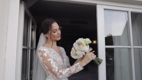 Mariée en robe de mariée et voile avec bouquet de fleurs de marié séjour près des portes du balcon à la maison — Video