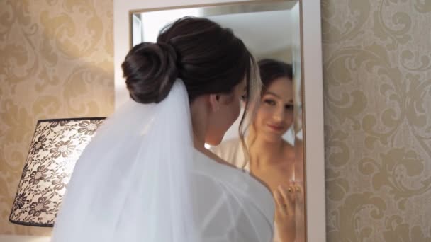 Novia en vestido de tocador blanco permanecer cerca del espejo, mirando a la reflexión, los preparativos de la boda en casa — Vídeos de Stock