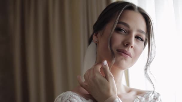 Bruid in witte jurk verblijf in de buurt van venster en aanraken van haar gezicht, bruiloft ochtend voorbereidingen thuis — Stockvideo