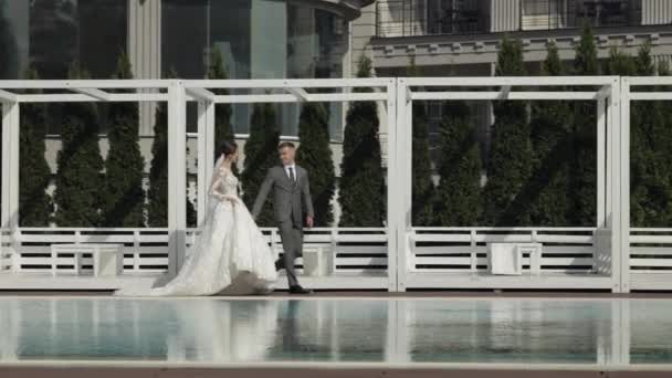 Recién casados novio novia caucásica caminando cerca de la piscina, cogidos de la mano, la familia de la pareja de boda — Vídeo de stock