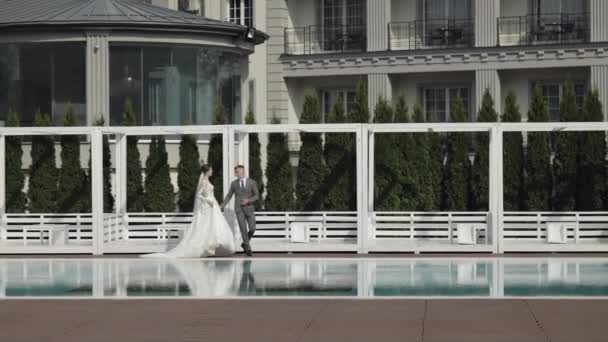 Recién casados novio novia caucásica caminando cerca de la piscina, cogidos de la mano, la familia de la pareja de boda — Vídeo de stock