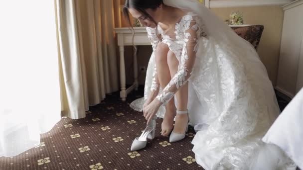 Jovem noiva em vestido de noiva branco vestindo sapatos de salto alto em pernas finas enquanto sentado em casa — Vídeo de Stock