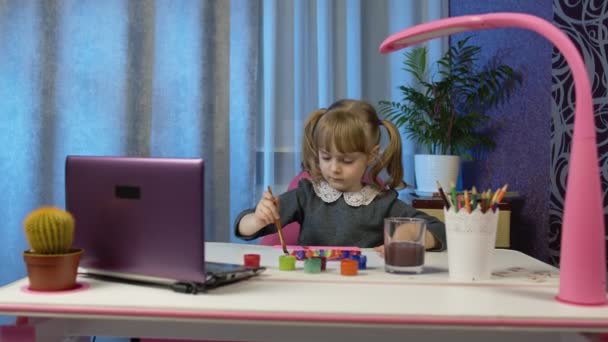 Kız evde dizüstü bilgisayar ve uzaktan eğitim kullanarak fırça ve boyalarla hevesle resim çiziyor. — Stok video