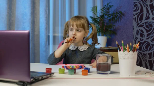 Девушка с энтузиазмом рисует кисточками и красками, используя ноутбук дома, дистанционное образование — стоковое фото