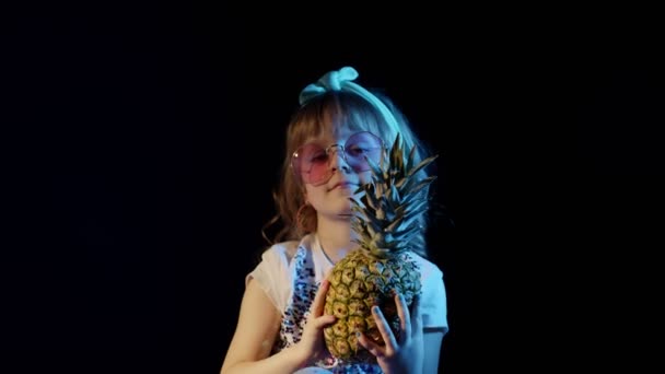 戴着未来派眼镜的漂亮女孩躲在菠萝后面，黑黑的背景上，笑着鬼鬼祟祟 — 图库视频影像