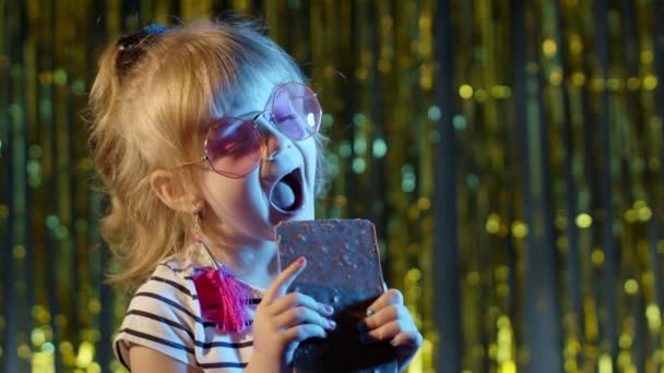 Stylowe modne małe dziecko dziewczyna na disco party cyberpunk klub jedzenie duży batonik czekolada słodkie cukierki — Wideo stockowe
