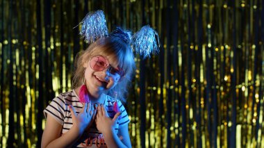 Modaya uygun bir kız pembe güneş gözlüğü takıyor, kameraya bakıyor, disko kulübünde neon ışıkta gülümsüyor.