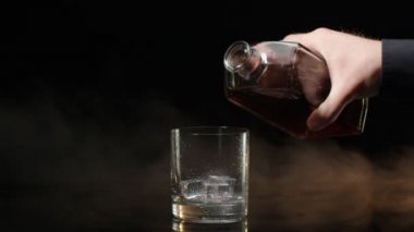 Barmen, viski konyağını şişeden bardağa doldurup, siyah arka planda buz küpleri olan bardağa doldurur.