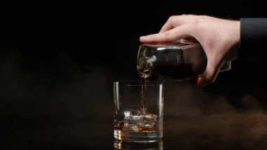 Barmen viski, konyak, şişeden cama konyak ve siyah arka planda buz küpleri dolduruyor.