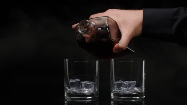 Bartender hälla whisky, konjak, konjak från flaska i glas med isbitar på svart bakgrund — Stockvideo