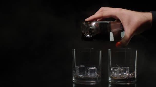 Barman pour golden whiskey cognac brandy from bottle into glasses with ice cubes on dark background — Stock Video