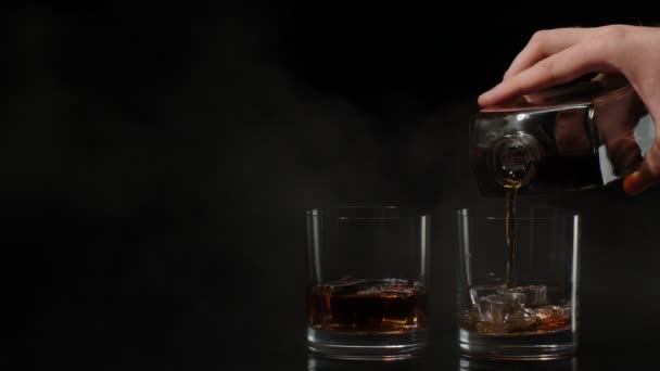 Barman schenkt whisky, cognac, cognac uit fles in glazen met ijsblokjes op zwarte achtergrond — Stockvideo