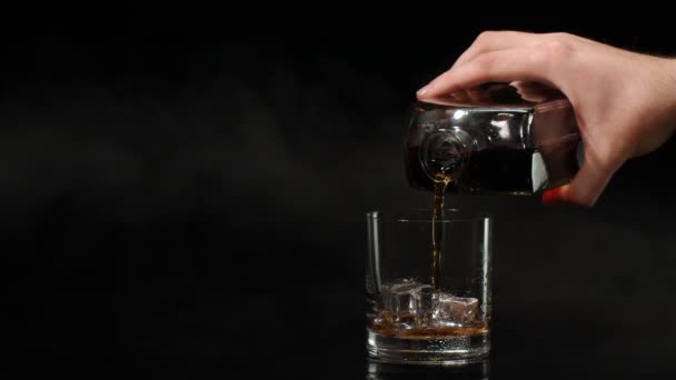 Barman pour whiskey cognac brandy from bottle into drinking glass with ice cubes on dark background — Stock Video