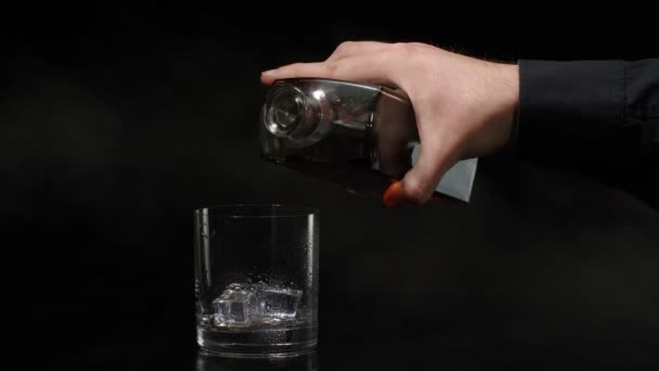 Barman schenkt whisky, cognac, cognac uit fles in glas met ijsblokjes op zwarte achtergrond — Stockvideo