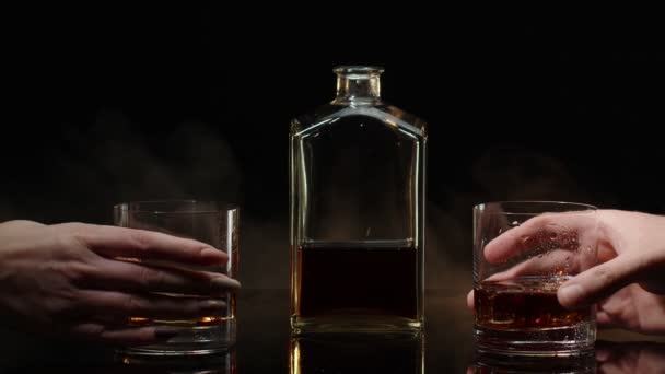 Twee handen met een glas cognac whisky met ijsblokjes proost op zwarte achtergrond — Stockvideo